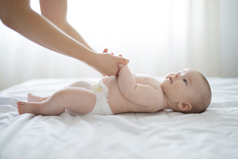 Zufriedenes Baby mit Windel liegt auf einem Bett und wird an den Händen gehalten