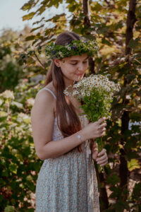 Junge Frau mit Kräuter-/Blumenkranz in den langen braunen Haaren und Kamillenblüten-Strauß in der Hand