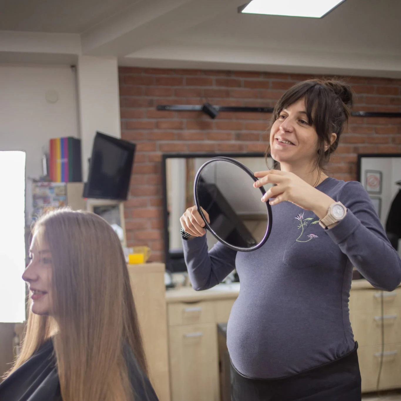 Schwangere Friseurin zeigt einer Kundin mit langen Haaren die Frisur mit einem Handspiegel im Salon.