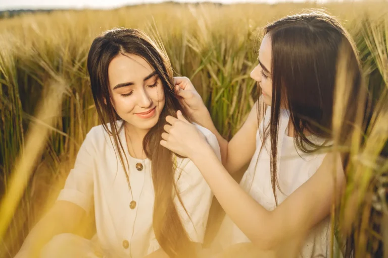 Zwei junge Frauen sitzen im goldenen Getreidefeld, eine berührt sanft das Haar der anderen – Symbol für Nähe und Redewendungen rund um Haar und Sprache.